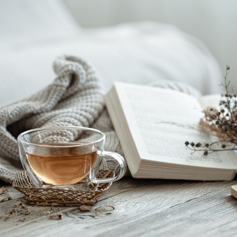 Tasse de thé chaud à côté d'un livre ouvert sur un plaid en maille, illustrant l'accompagnement des futurs parents en Normandie par Parenthée.